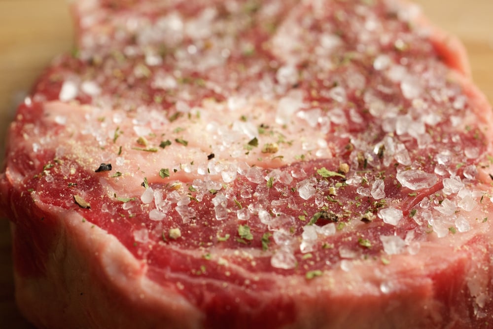 A raw steak covered in coarse salt and chopped dried herbs