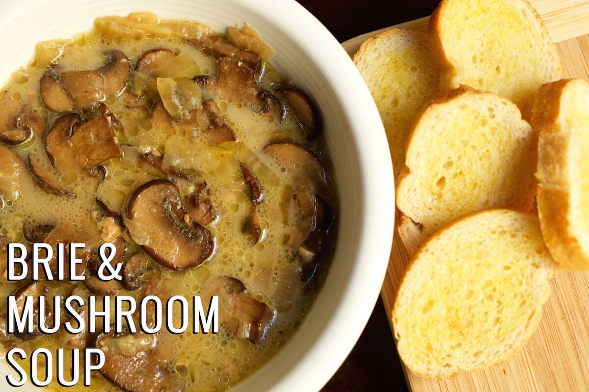 Mushroom soup in a white bowl next to a cutting board with sliced baguette. Text in the left hand corner reads "Brie & Mushroom Soup".