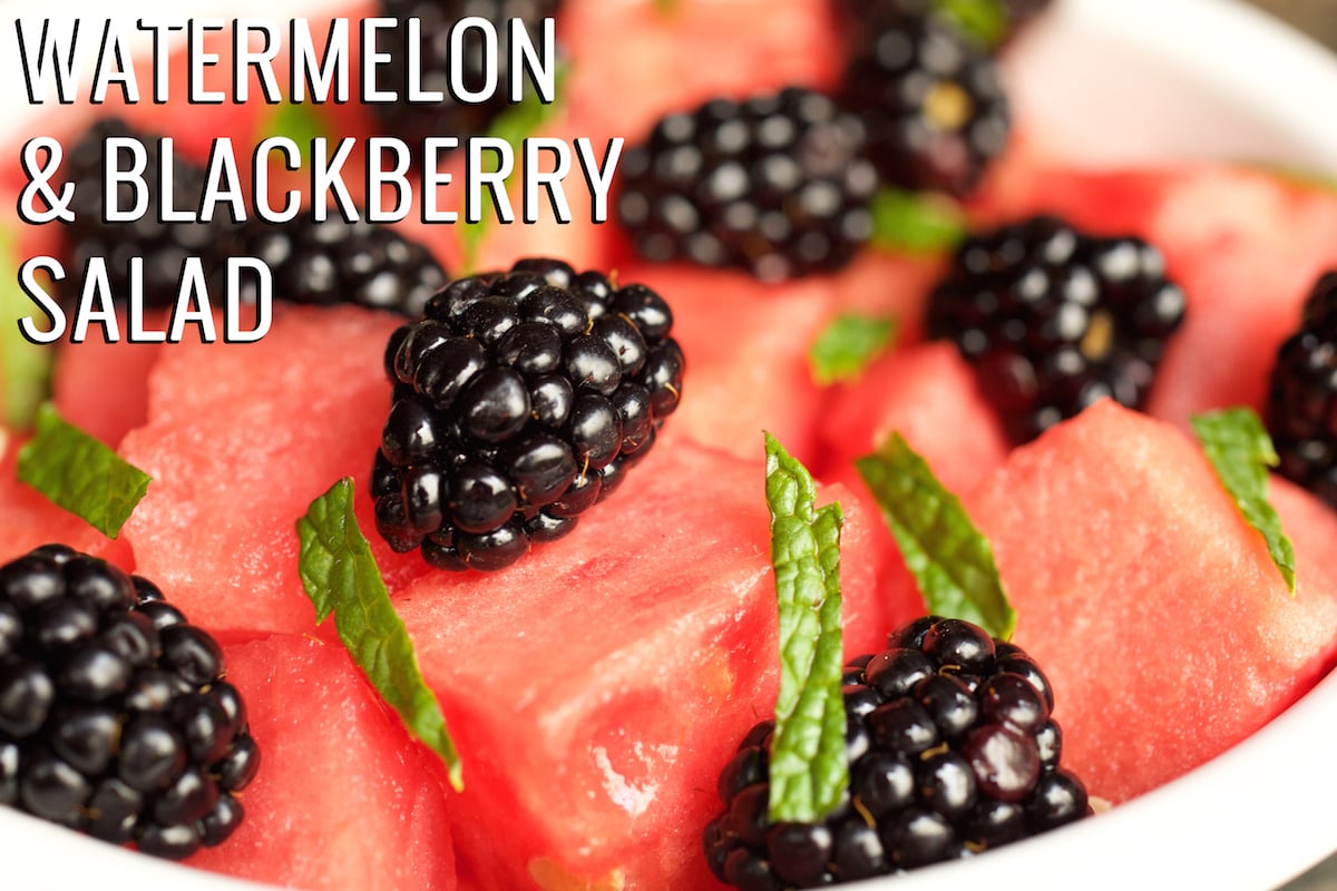 A close up of a bowl with cut watermelon, whole blackberries, and chopped mint. Text in the upper left hand corner reads "Watermelon & Blackberry Fruit Salad"