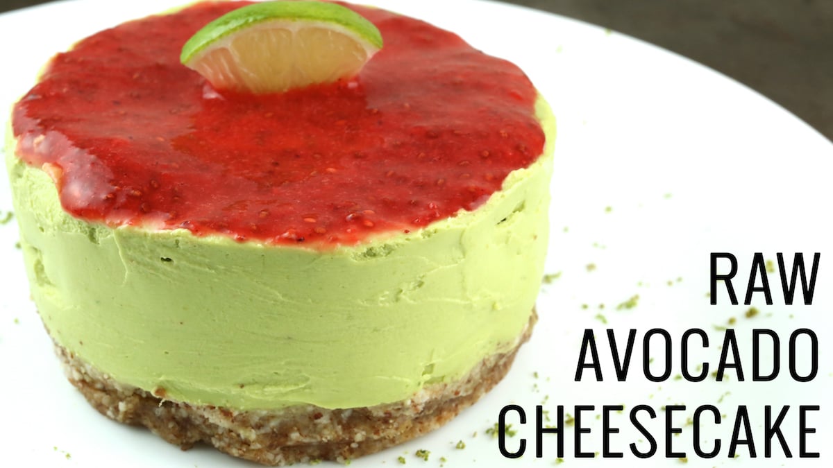 A small, green cheesecake with raspberry puree topping that is garnished with a lime. Text reads "Raw Key Lime Avocado Cheesecake"