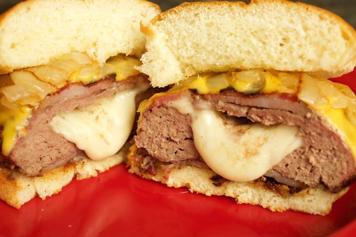 A bacon wrapped cheese stuffed burger sliced in half to show the cheese oozing out of the inside.