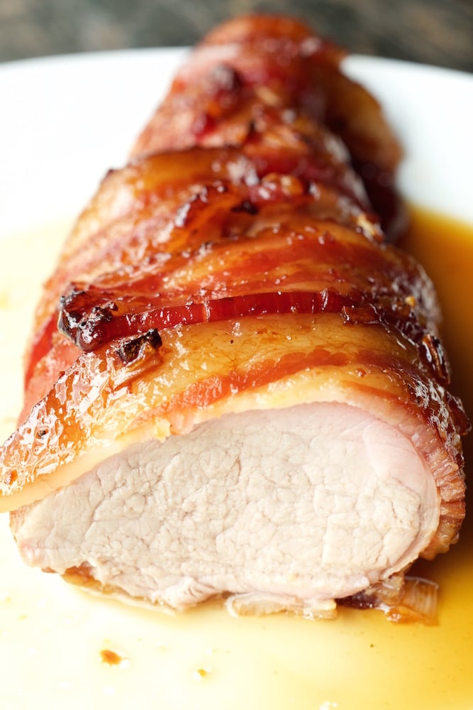 Close up of a sliced bacon wrapped pork tenderloin sitting on a white plate in a pool of it's own juices.