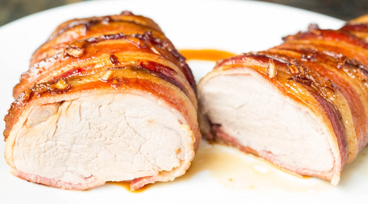 A pork tenderloin that's been wrapped all the way around with bacon has been sliced in half to show the perfectly cooked inside.