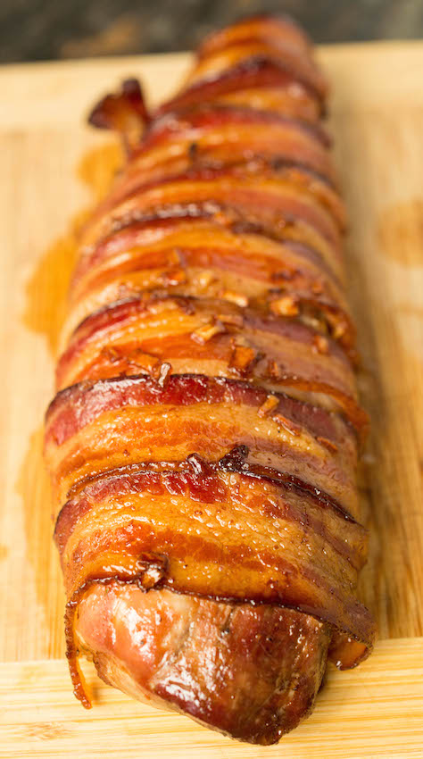 A bacon wrapped pork tenderloin resting on a cutting board after it has been cooked.