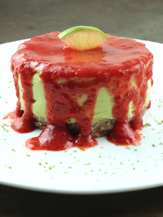 A small avocado cheesecake topped with raspberry puree and a lime slice sitting on a white plate.