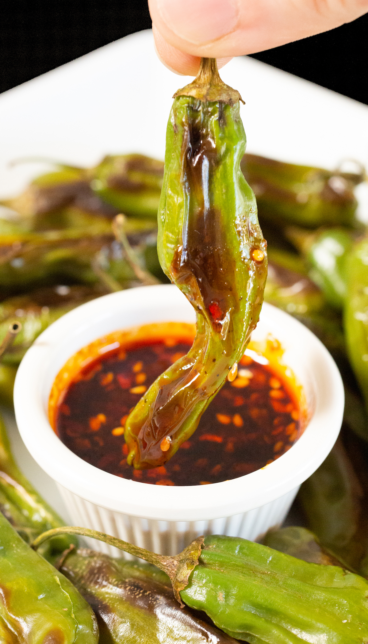 Two fingers hold a shishito pepper by the stem after it has been dipped in sauce.