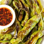 Blistered shishito peppers on a white plate next to a small container of brownish red dipping sauce.