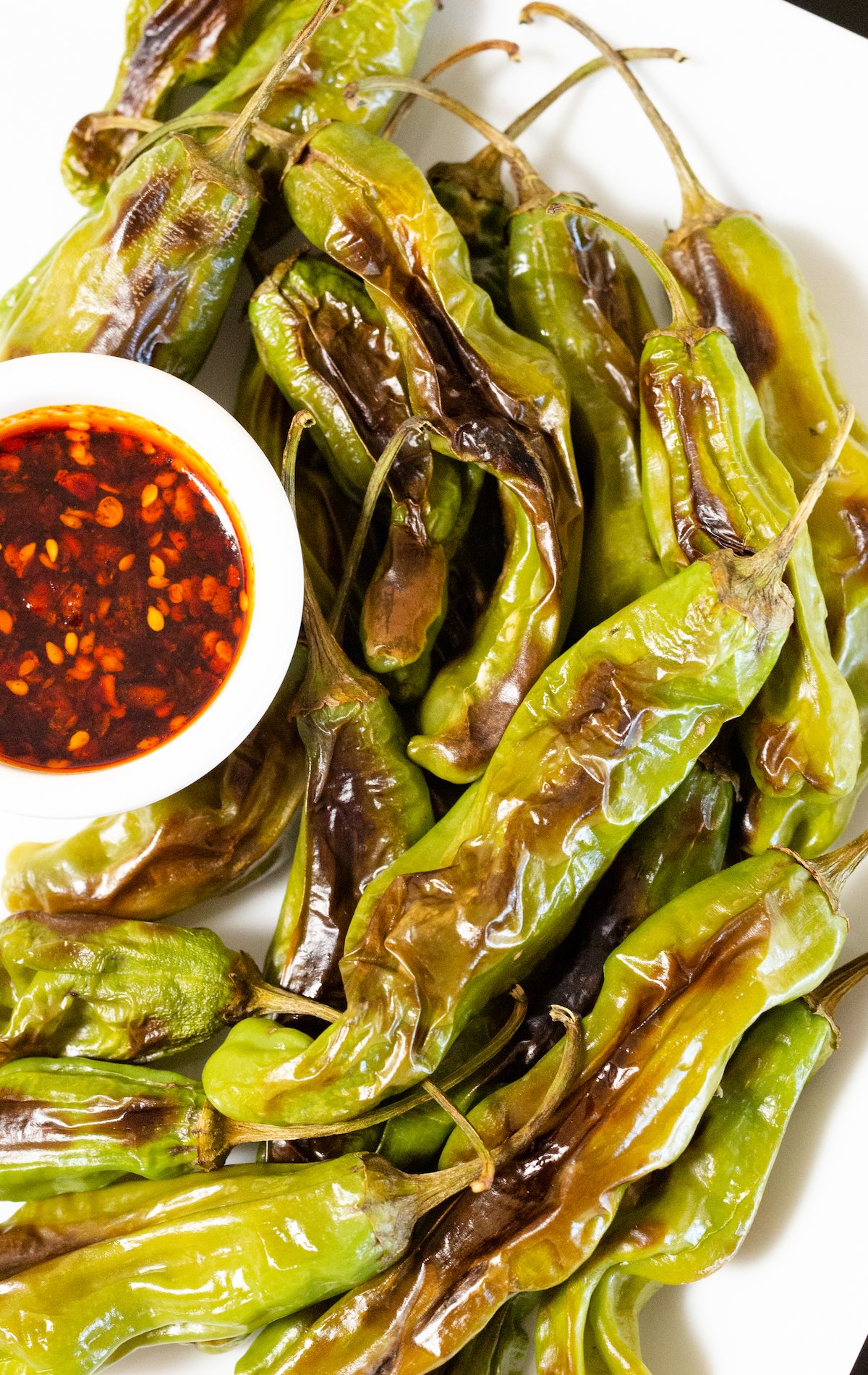 Blistered shishito peppers on a white plate next to a small container of brownish red dipping sauce.