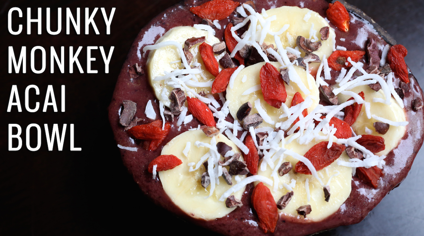 A coconut shell bowl is filled to the brim with purple acai mixture and topped with sliced bananas, cacao nibs, goji berries, and shredded coconut. Text to the left reads "Chunky Monkey Acai Bowl"