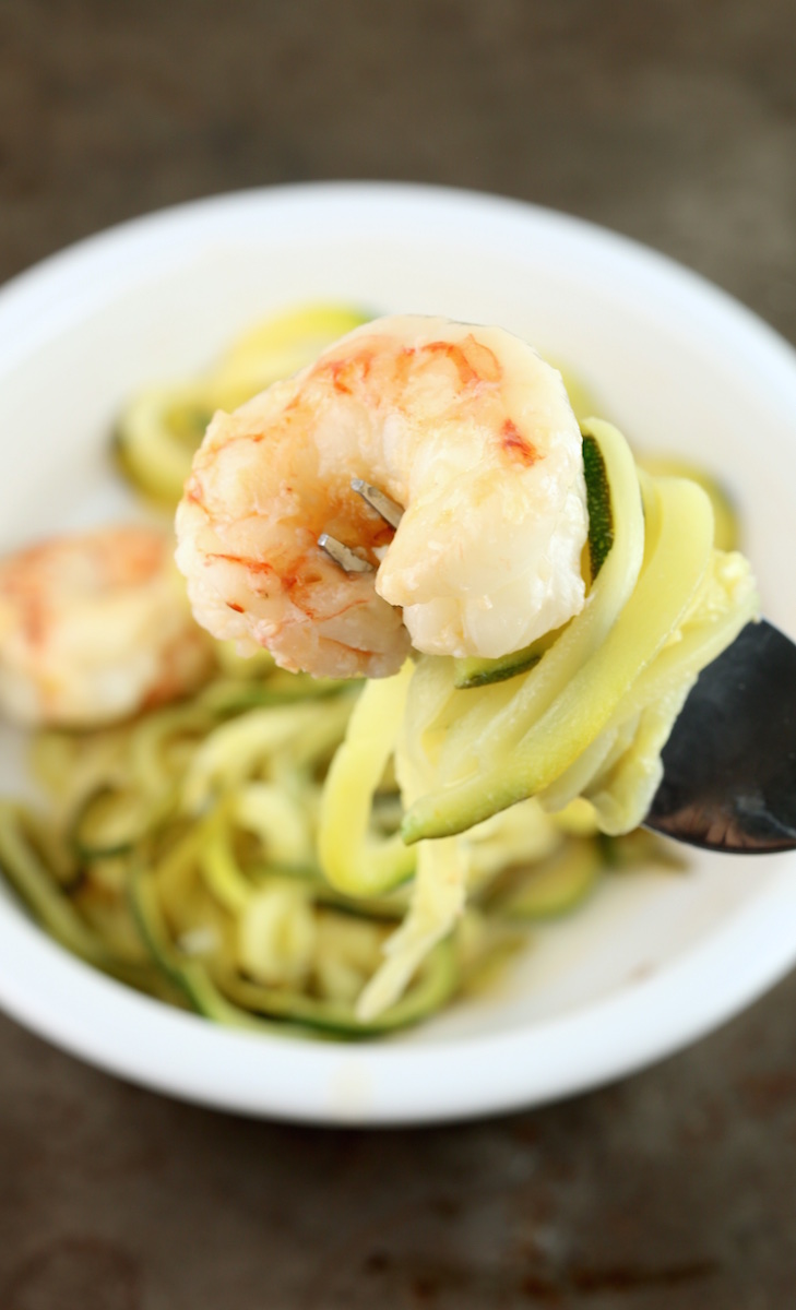 A fork holds zucchini noodles and a single shrimp over a white bowl filled with zucchini shrimp scampi.