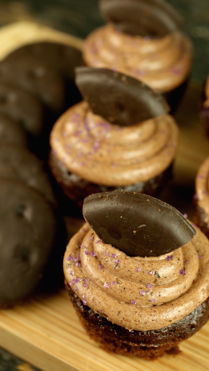 Several small chocolate cupcakes that are garnished with thin mint cookies sit on a cutting board.