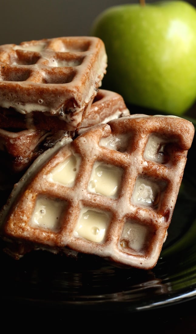 An Apple Fritter Waffle Donut dripping with glaze. More are stacked in the background next to a green apple.