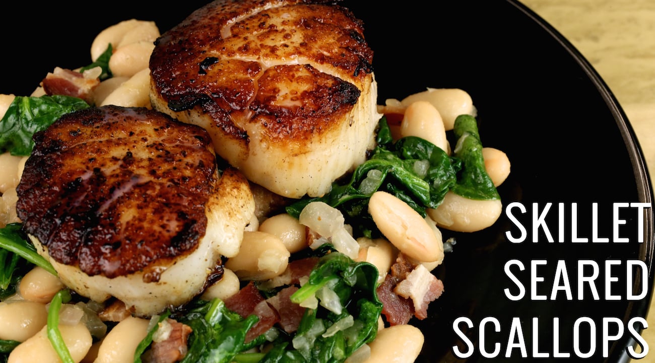A black dinner plate with two large seared scallops sitting on a bed of white beans, spinach, and bacon.