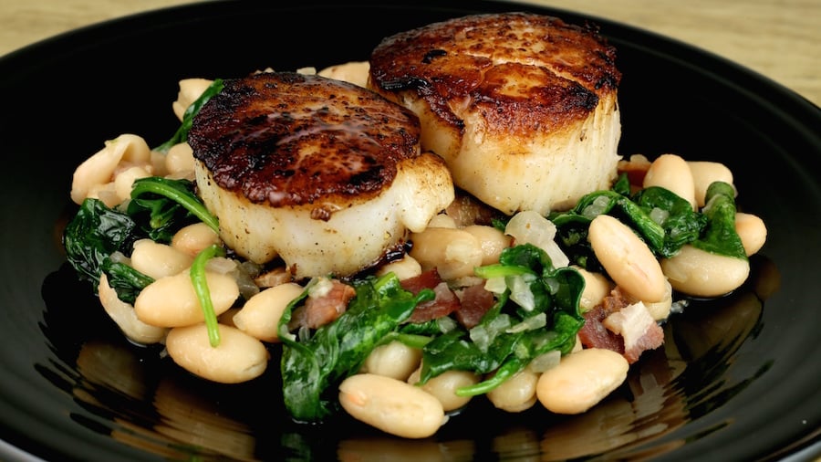 A black dinner plate with two large seared scallops with white beans & spinach