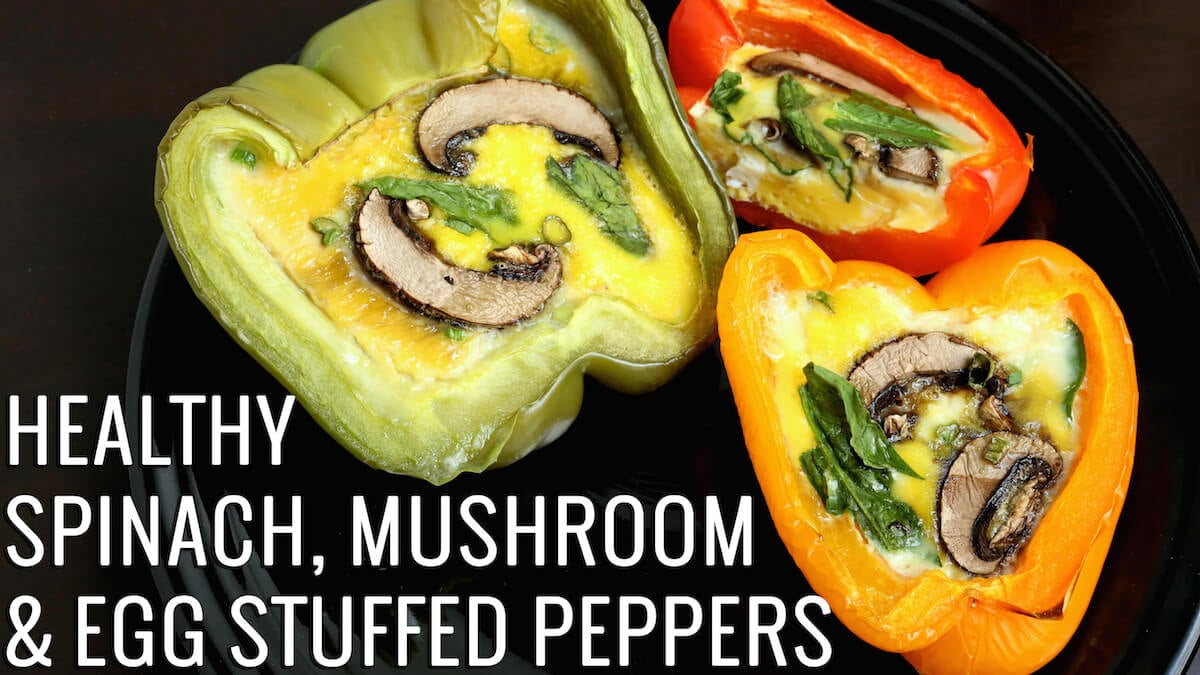 Red, Green, and orange bell peppers that have been sliced in half and baked with eggs, vegetables, and mushrooms sit on a black plate. Text reads: "Healthy Spinach, Mushroom, & Egg Stuffed Peppers"