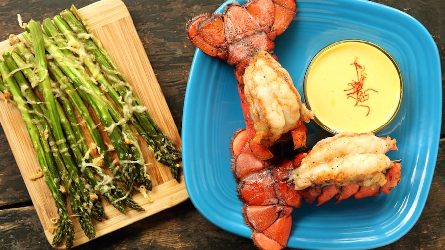 A plate with two broiled lobster tails and a bowl of aioli. A small cutting board with baked asparagus sits next to it.