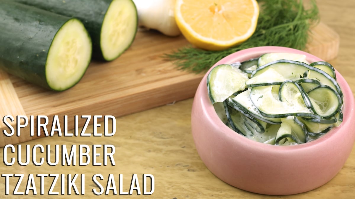 A pink bowl of Spiralized Cucumber Tzatziki Salad next to sliced cucumbers, lemon, dill, and garlic on a cutting board.