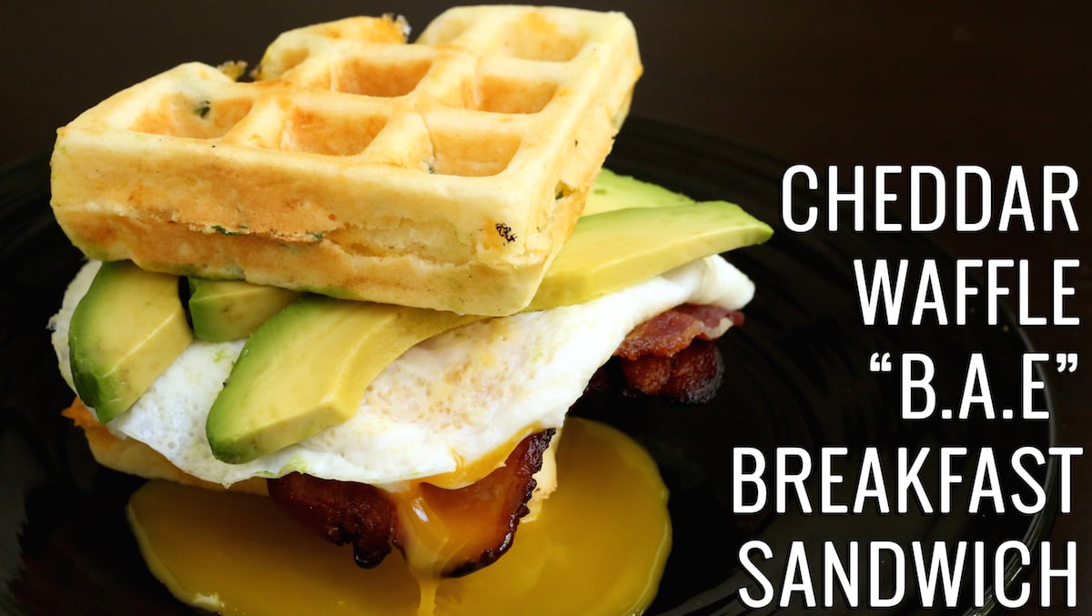 A breakfast sandwich featuring slices of bacon, a fried egg, and sliced avocado on two waffles. Text reads "Cheddar Waffle BAE Breakfast Sandwich"