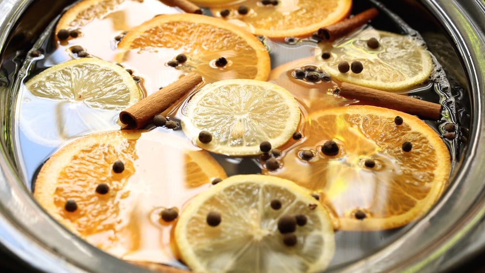 Overhead view of apple cider cooking in the crock pot. Slices of oranges and lemons are floating on top as well as allspice berries and three cinnamon sticks.