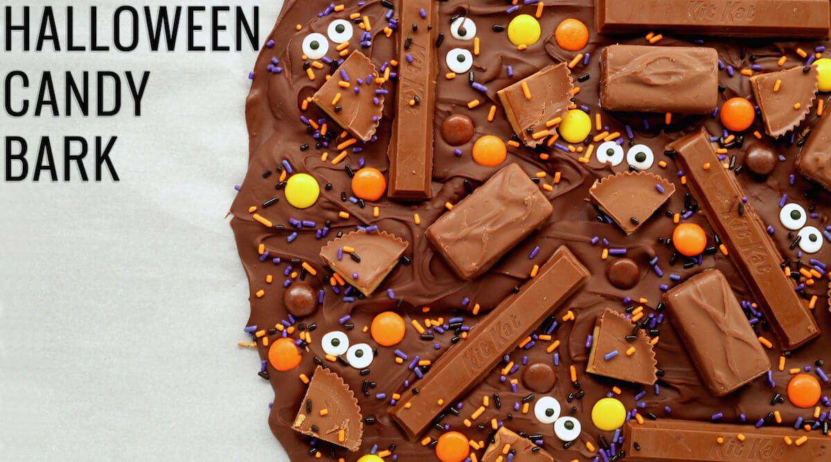 A close up of melted chocolate with mini Halloween candies and edible eyes on parchment paper. Text reads "Halloween Candy Bark"