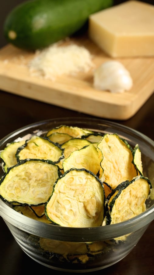 A small glass bowl of Zucchini Chips sits in front of a cutting board with the ingredients (raw zucchini, shredded parmesan, and garlic).