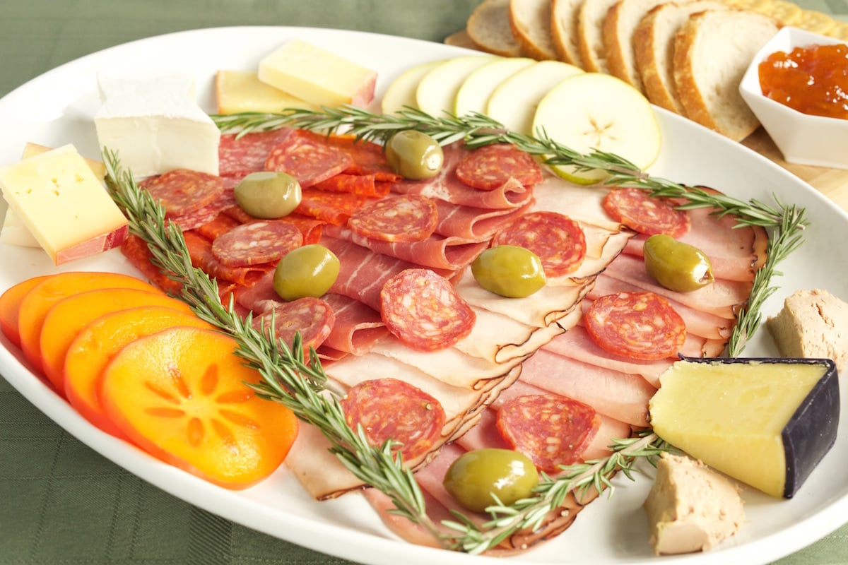 Meats and cheeses arranged on a large white platter in the shape of a Christmas Tree. Olives are used as ornaments, a block of white cheddar for the trunk, and brie cut into a star for the topper.