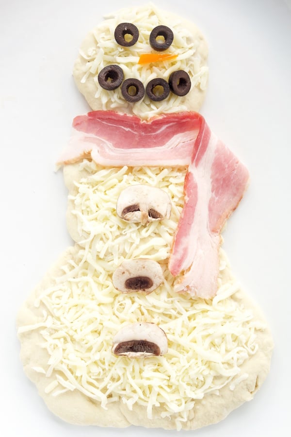 Pizza dough in the shape of a snowman, topped with cheese and mushroom slices for bottoms, a slice of bacon for the scarf, black olives for the eyes and mouth, and a nose made out of a small triangle carrot slice.