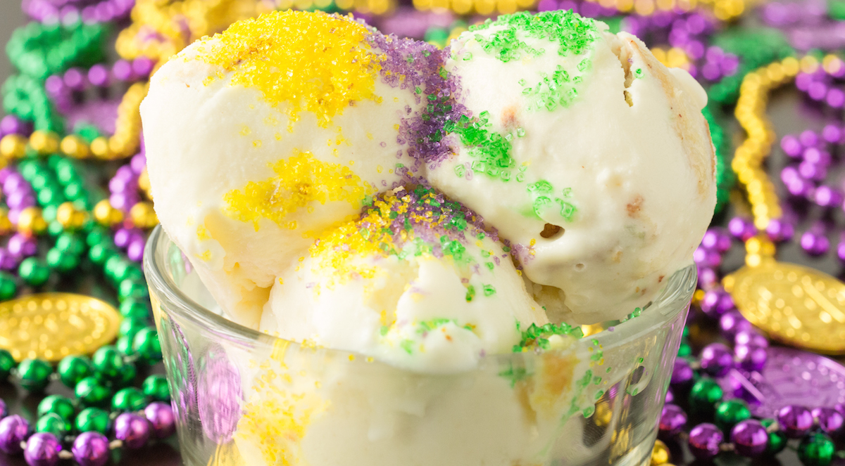 Three scoops of King Cake ice cream in a glass bowl that's surrounded by Mardi Gras beads. This scoops are topped with yellow, purple, and green sanding sugar.