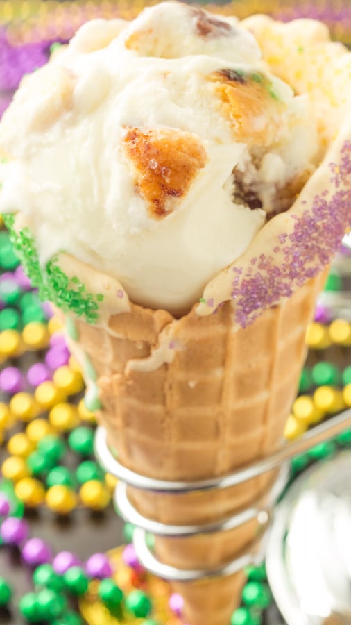 A waffle cone filled with king cake ice cream. Mardi gras beads are in the background out of focus.