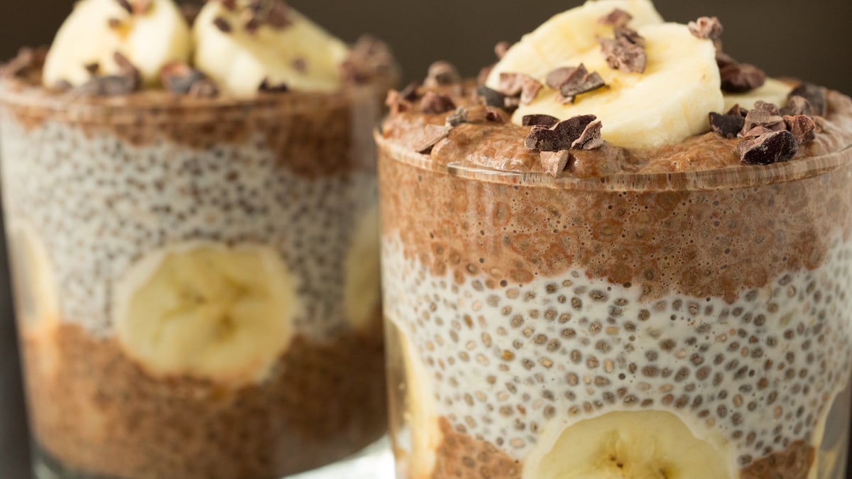 Two lowball glasses are filled with layered banana chocolate chia pudding that's topped with sliced banana and cacao nibs.