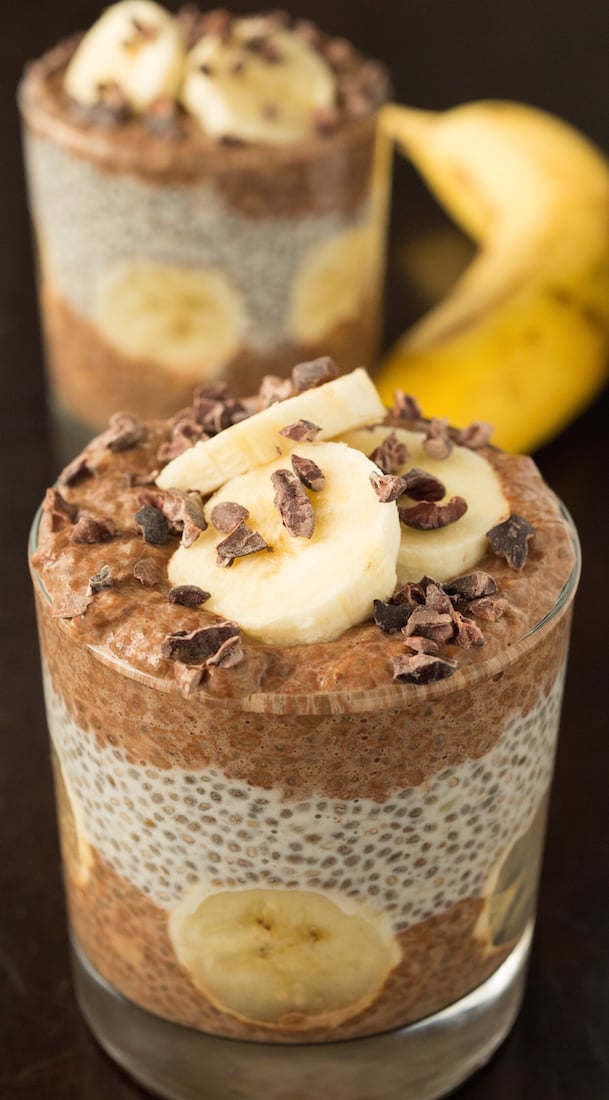 In focus in the foreground is a glass filled with layered chocolate and banana chia pudding with fresh banana slices and cacao nibs on top. Out of focus in the background is another glass and a whole banana.