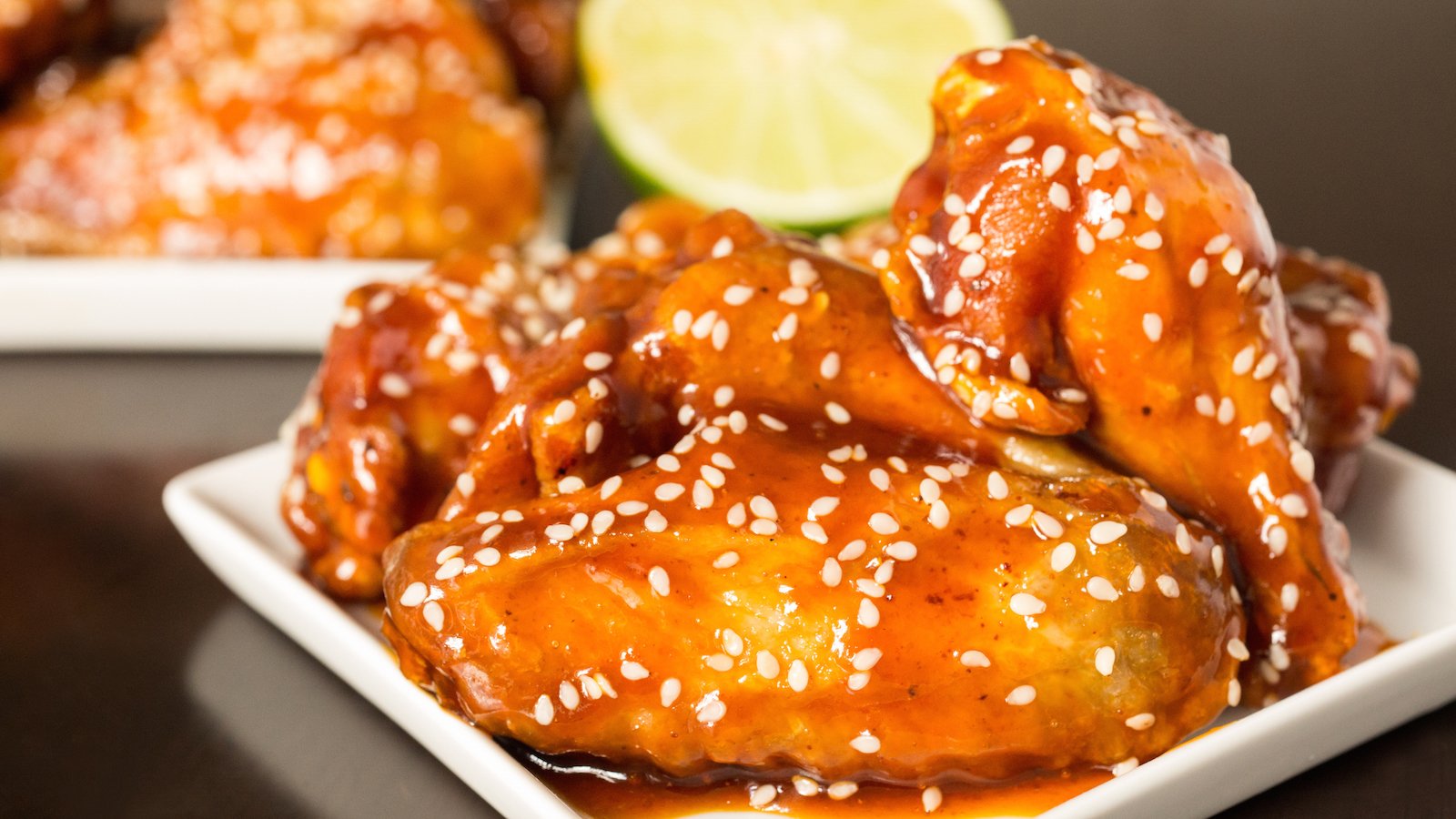 In focus in the foreground is a small appetizer plate with chicken wings covered in a bright orange honey sriracha lime wings. They are garnished with sesame seeds and a half lime sits out of focus in the background along with another small plate of wings. 