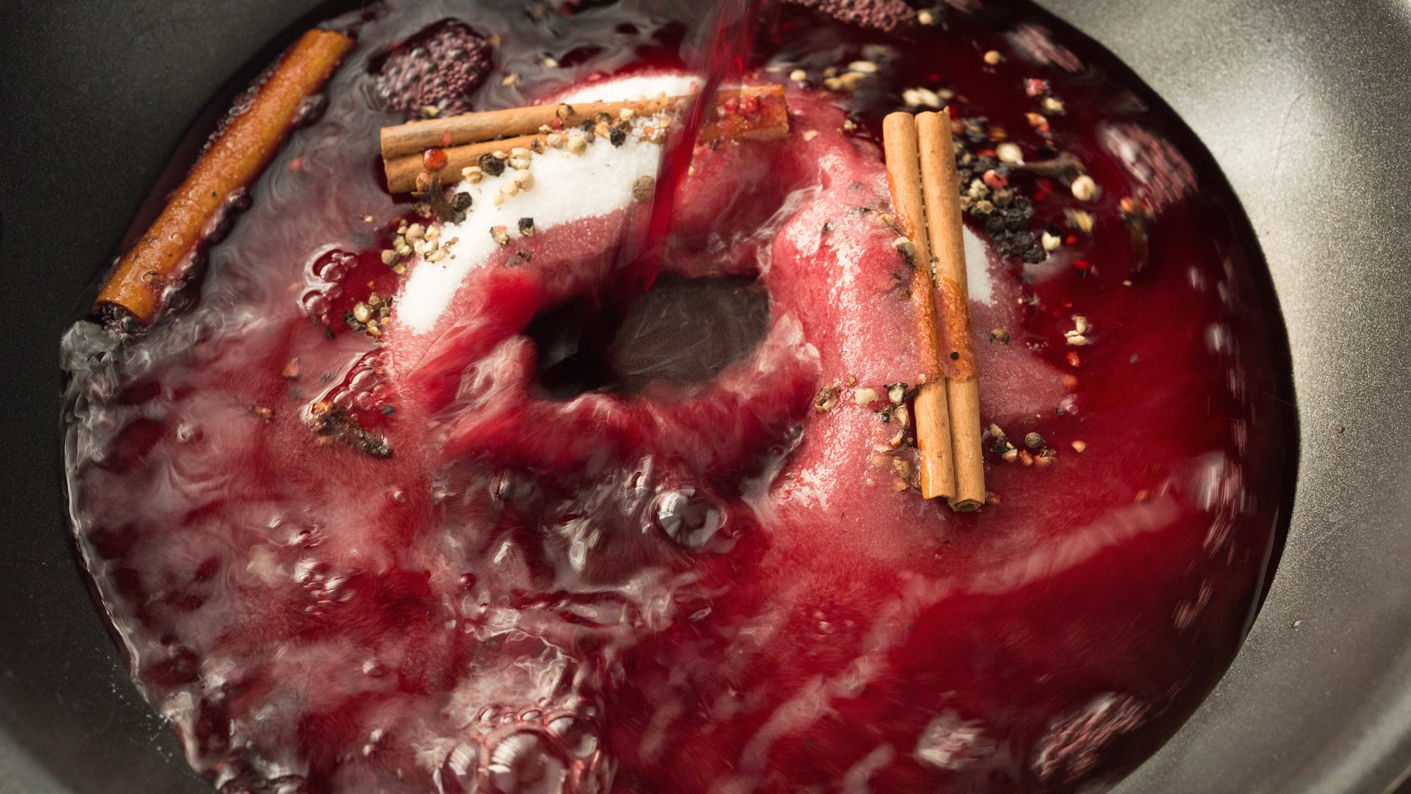 Sugar, cinnamon sticks, and spices in a skillet with red wine being poured over to make Spiced Red Wine Syrup