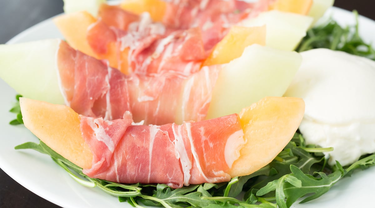 Alternating slices of cantaloupe and honeydew melon that are wrapped with prosciutto. They are sitting on a bed of arugula next to a ball of burrata.