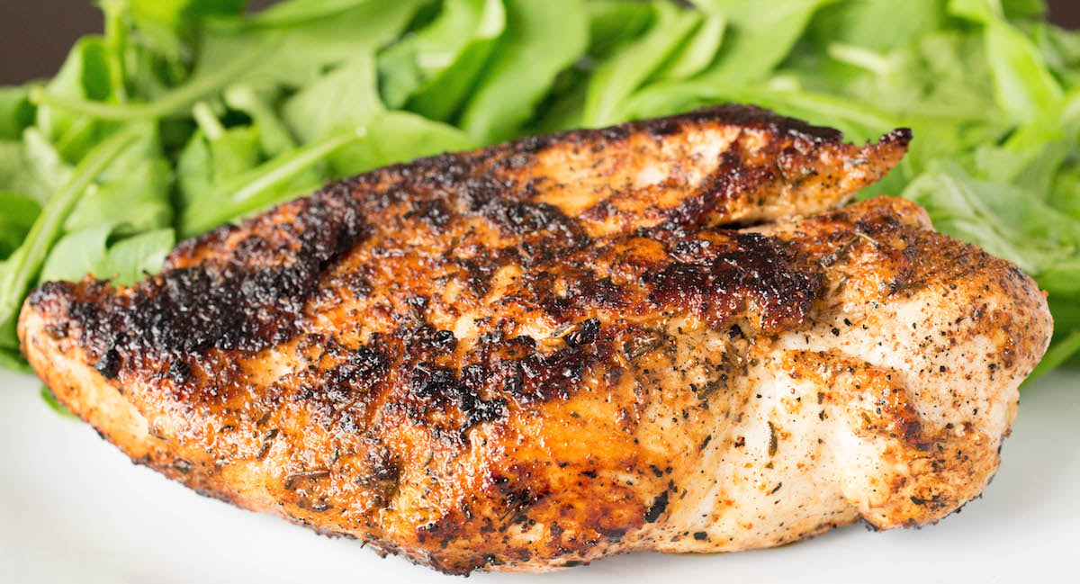 Horizontal few of a plump chicken breast that's been covered in blackening seasoning. A bed of greens sits out of focus in the background.