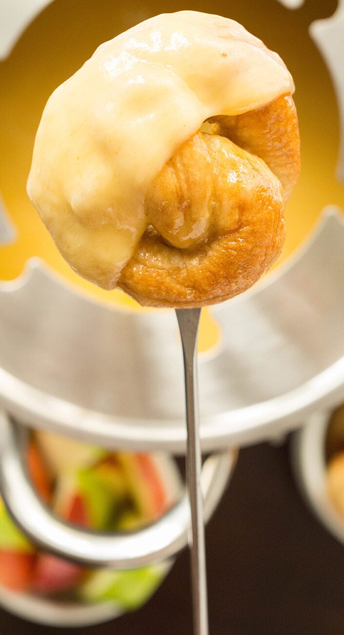 A mini soft pretzel that's been dipped in yellow fondue beer cheese is on a fondue skewer, the pot of fondue cheese is out of focus in the background.