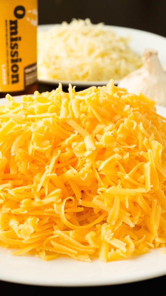 A plate filled with a pile of freshly shredded cheddar cheese is in focus in the foreground, a bottle of beer, a head of garlic, and a plate of shredded gruyere cheese is out of focus in the background.