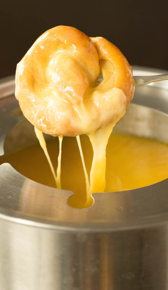 A fondue pot filled with beer cheese has a mini soft pretzel being dipped in.