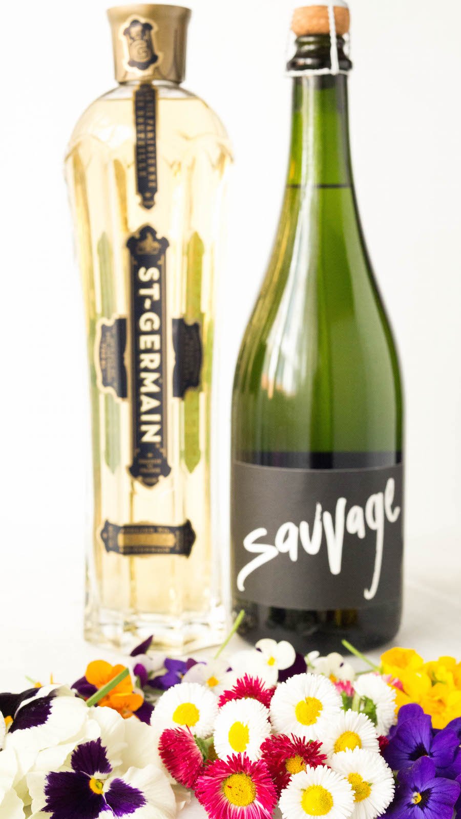 A bottle of St. Germain liqueur and "savage" champagne sit on a white background in front of a colorful assortment of edible flowers