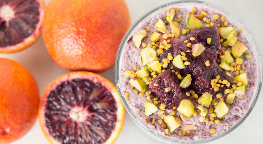 Overhead view of a small glass of Blood Orange Chia Seed layered with Vanilla Overnight Oats & topped with pistachios and bee pollen. Sliced blood oranges are out of focus in the background.