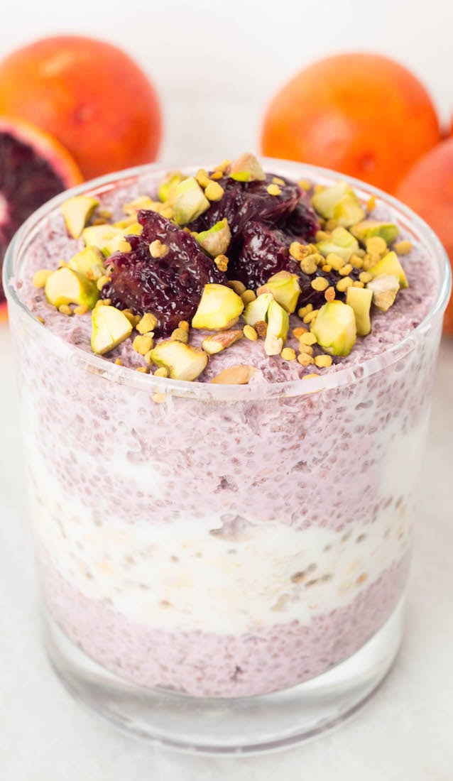 A lowball glass is filled with a layer of pink blood orange chia pudding, then a layer of white vanilla overnight oats, and another layer of the chia pudding on top. Chopped pistachios, blood orange, and be pollen garnish the top. Blood oranges are out of focus in the background.