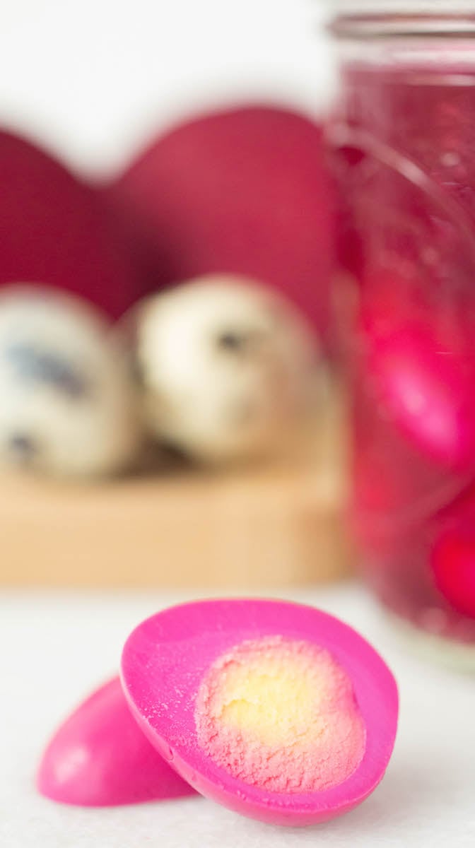 A boiled quail egg that has been turned pink by pickling it with beet juice has been sliced in half to show the yellow yolk. Out of focus in the background there are raw quail eggs, sliced beet, and a mason jar of beet juice. 