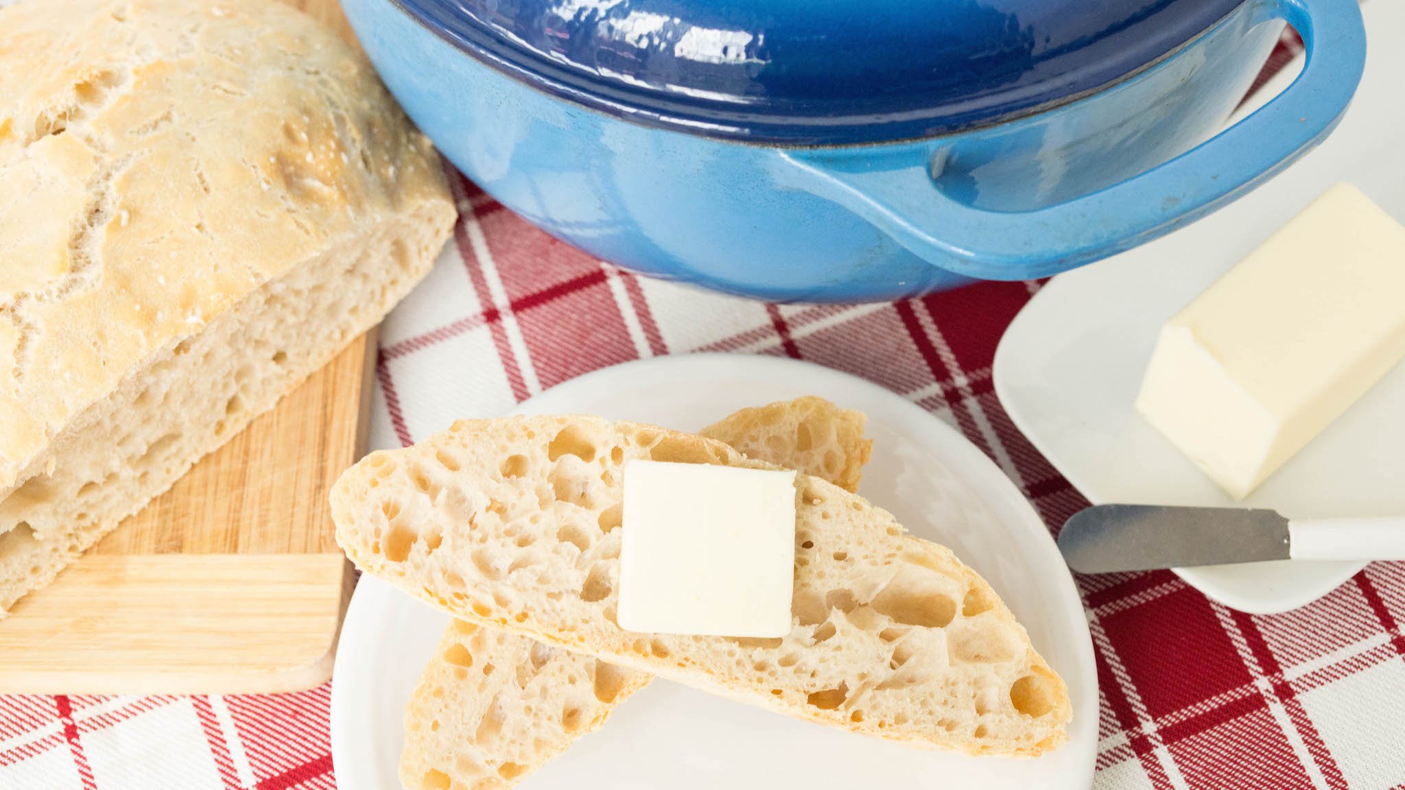 Overhead view of two slices of no knead dutch oven bread on a white plate next to the rest of the uncut loaf and the pot.