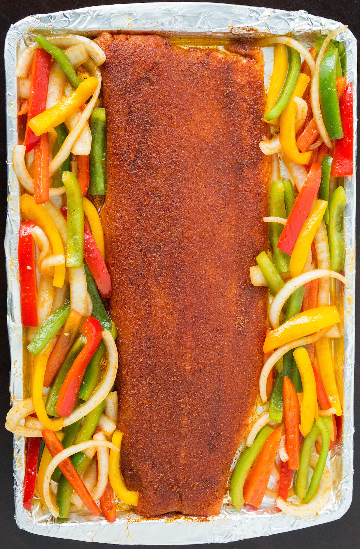 Overhead view of a large salmon fillet and sliced multi colored bell peppers and onions on a sheet pan