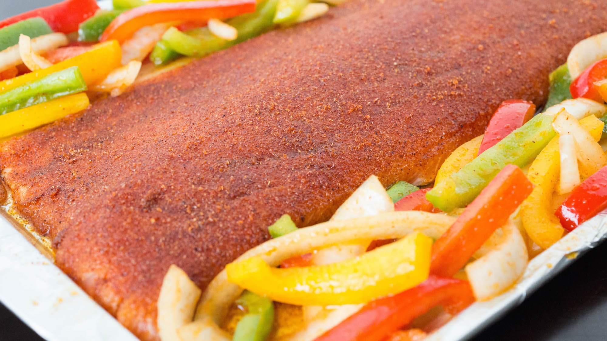 A sheet pan has been wrapped in aluminum foil. On top is a large salmon fillet that's been covered in fajita seasoning. Slice bell peppers and onions are on both sides. 