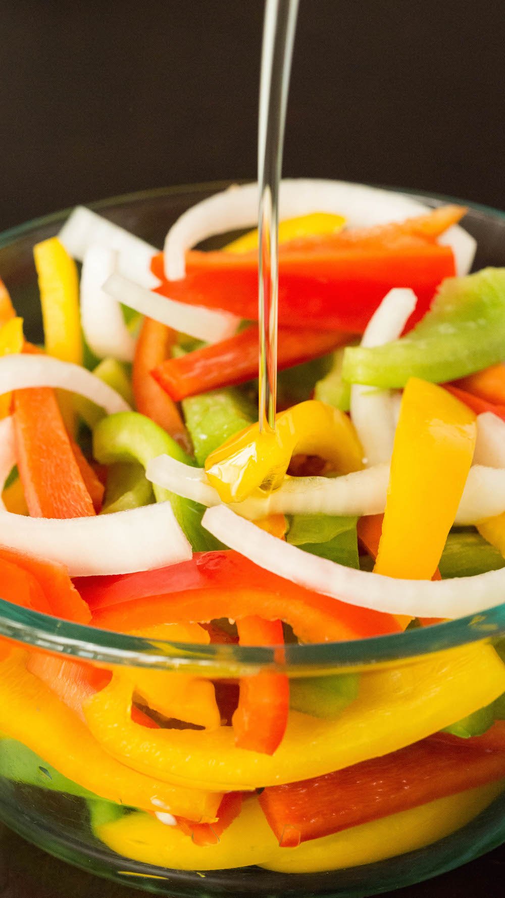 A large glass pyrex bowl filled with sliced green, red, and yellow bell peppers as well as white onions. Oil is being poured over the top.