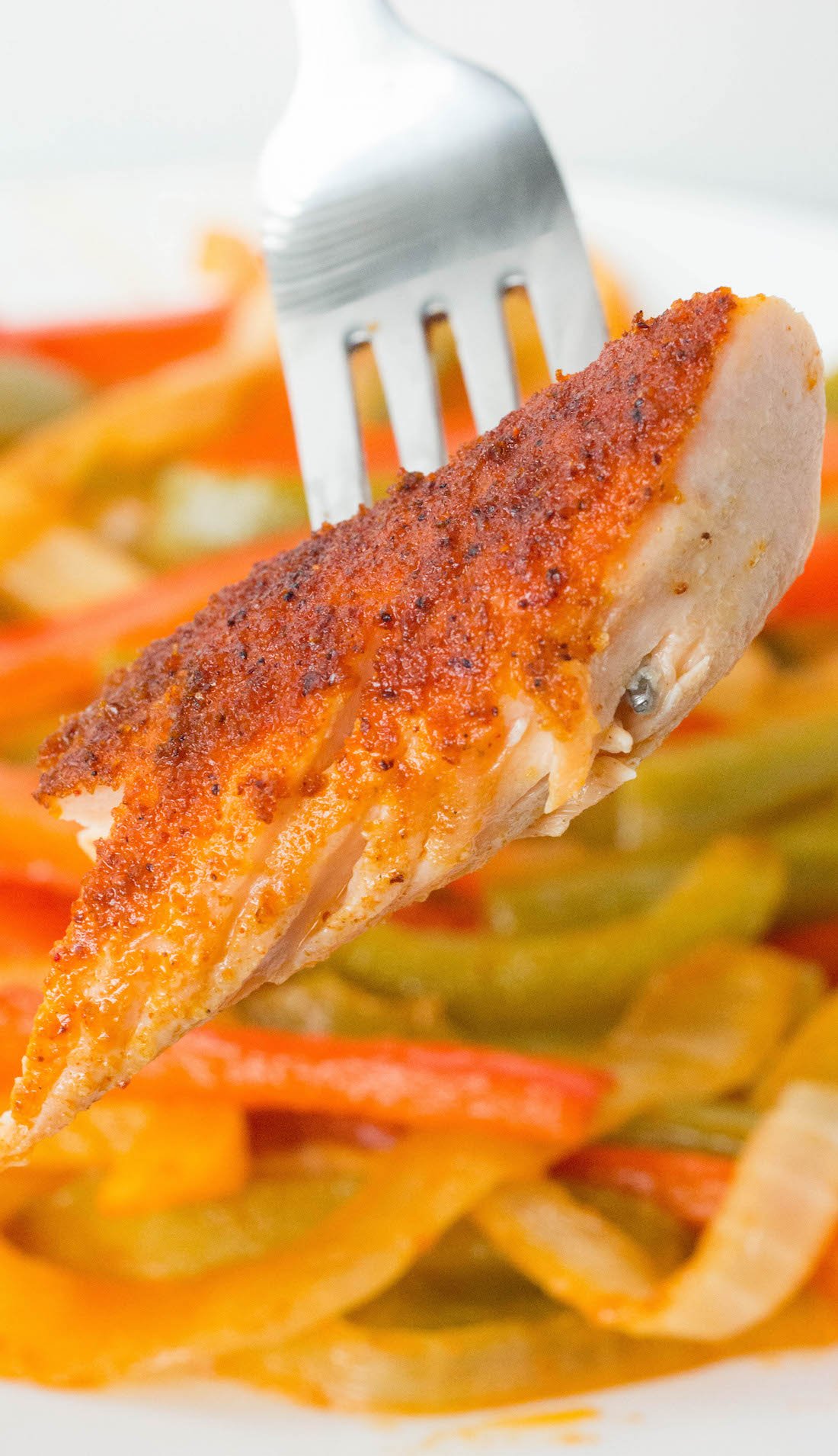 Close up of a piece of a fajita seasoned salmon on a fork. Cooked sliced bell peppers and onions are out of focus in the background.