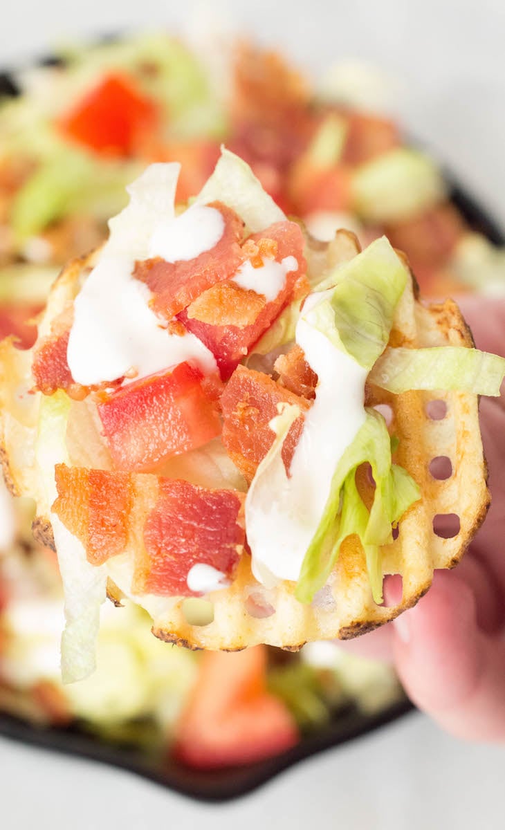 Close up of a hand holding up a BLT waffle fry (a waffle fry covered in melted mozzarella, chopped tomato, chopped bacon, sliced lettuce, and ranch dressing.