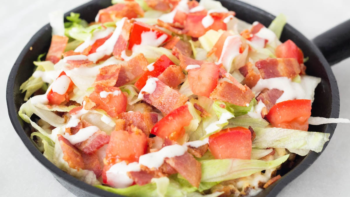 A cast iron skillet has been filled with waffle fries on the bottom, then topped with melted mozzarella cheese, slice lettuce, chopped tomatoes, chopped bacon and ranch dressing to make BLT waffle fries.