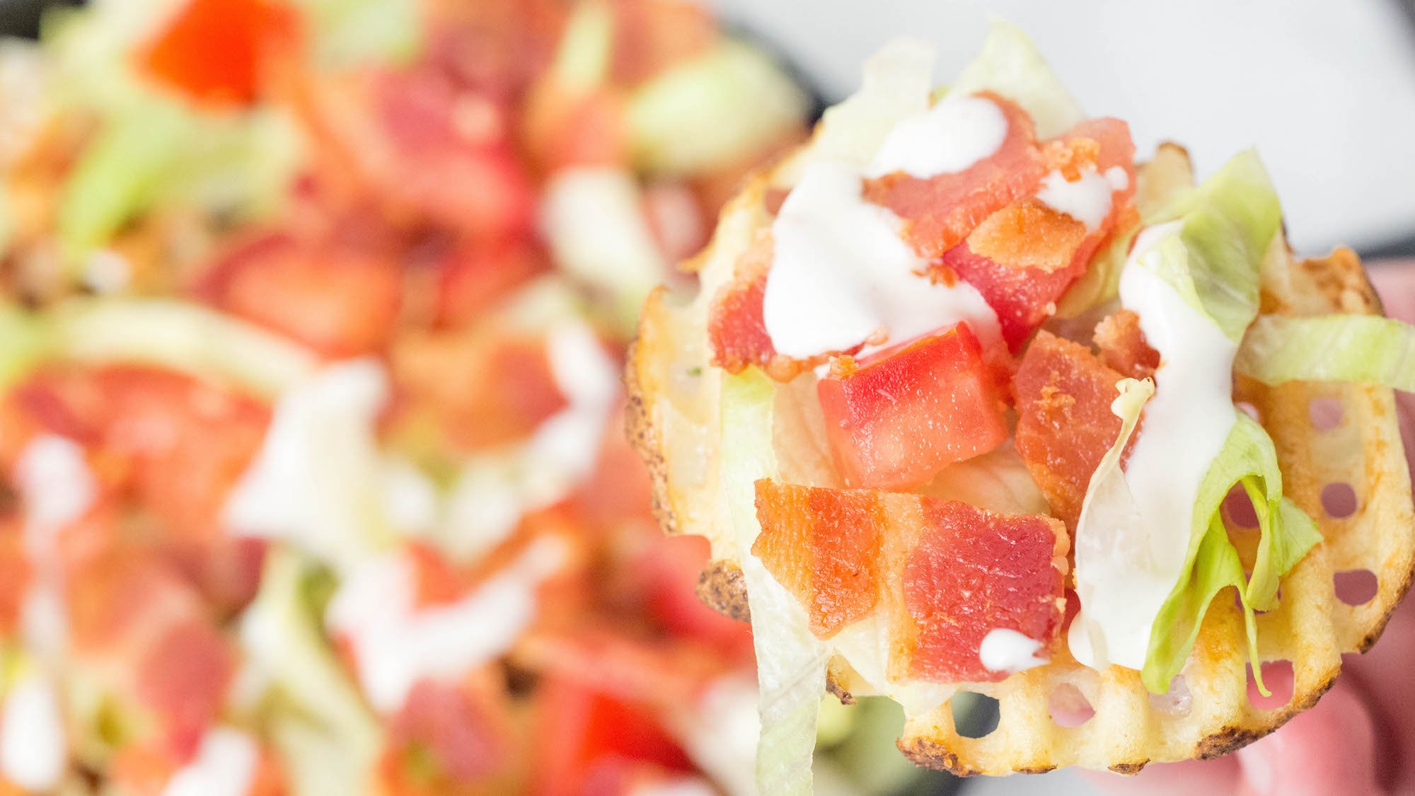 A hand holds up a waffle fry that's been covered in sliced lettuce, chopped tomatoes, chopped bacon, then drizzled in ranch. More BLT fries are out of focus in the background.